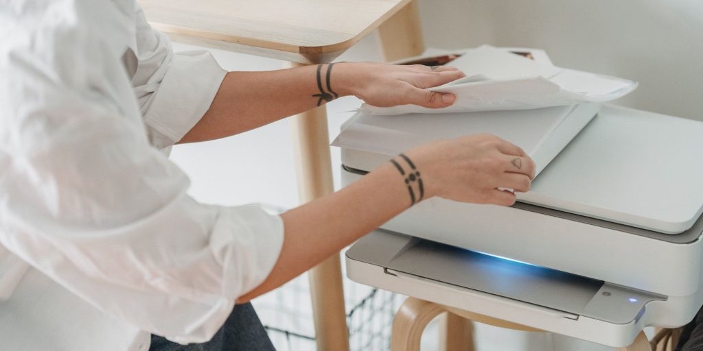 A person moves papers on top of a printer with its lights turned on